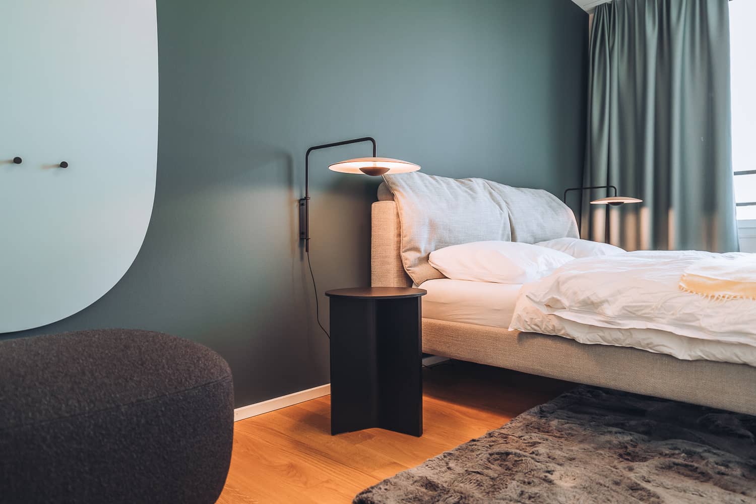Neben einem Bett ist eine Nachttischlampe an der Wand angebracht, welche den Nachttisch beleuchtet